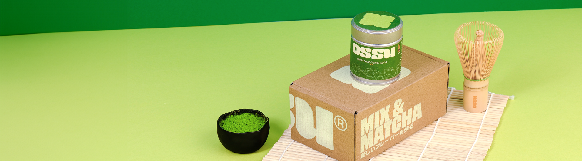 OSSU Golden Grade organic matcha tin on a cardboard box with a bowl of matcha powder and bamboo whisk on a green background