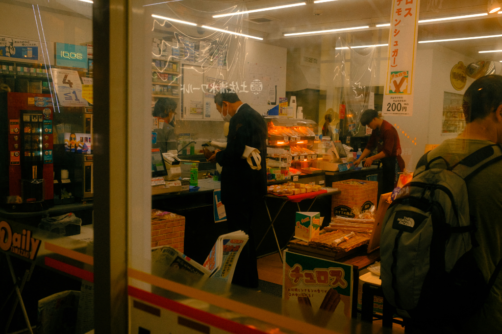 Interior of a small convenience store in Japan with customers paying at the counter and shelves of snacks and drinks under bright lights