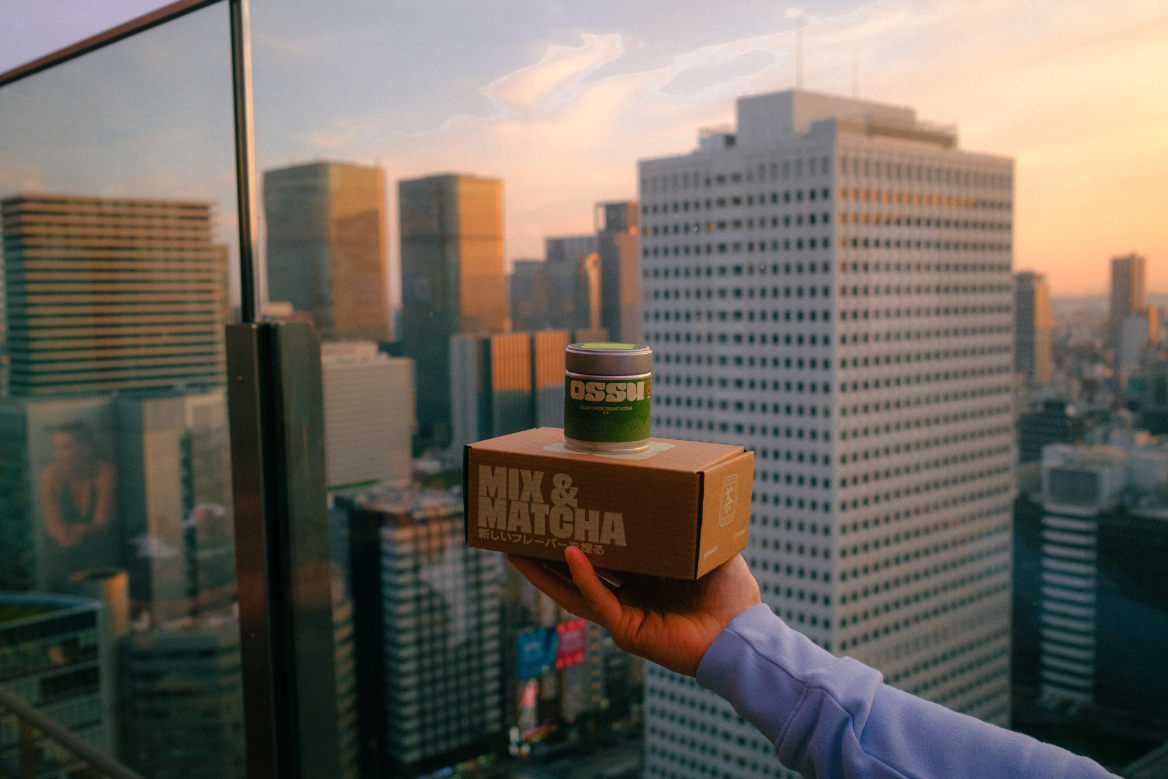 Hand holding an OSSU matcha tin and box overlooking a city skyline at sunset