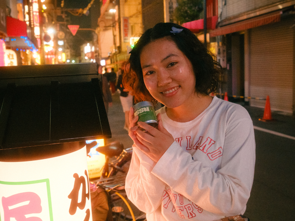 Smiling woman holding an OSSU matcha tin on a lively Japanese street at night