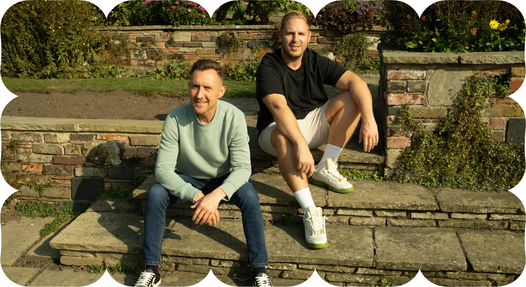 Two men sitting on stone steps outdoors in a garden setting, smiling in sunlight