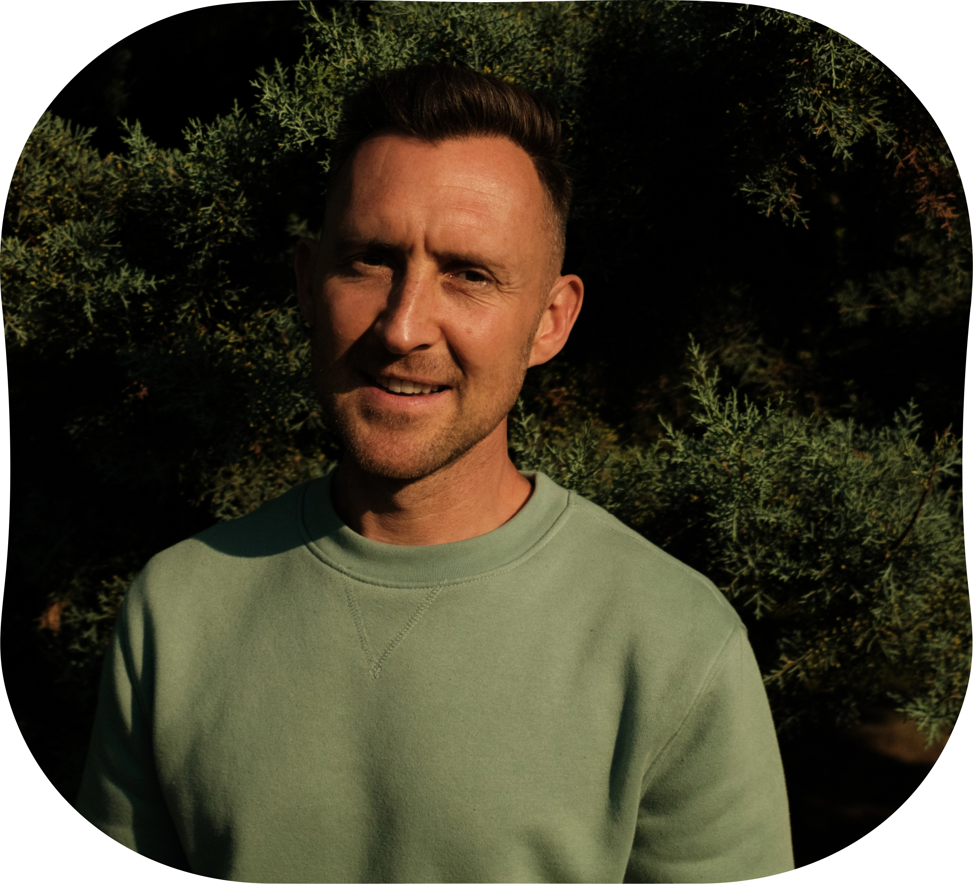 Man standing outdoors in sunlight, wearing a green sweatshirt with leafy trees in the background.