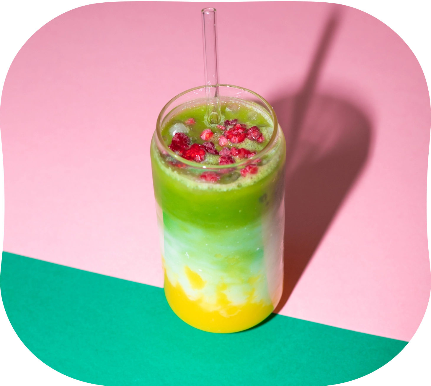 Layered iced mango matcha drink topped with crushed raspberries in a tall glass with a straw on a pink and green background