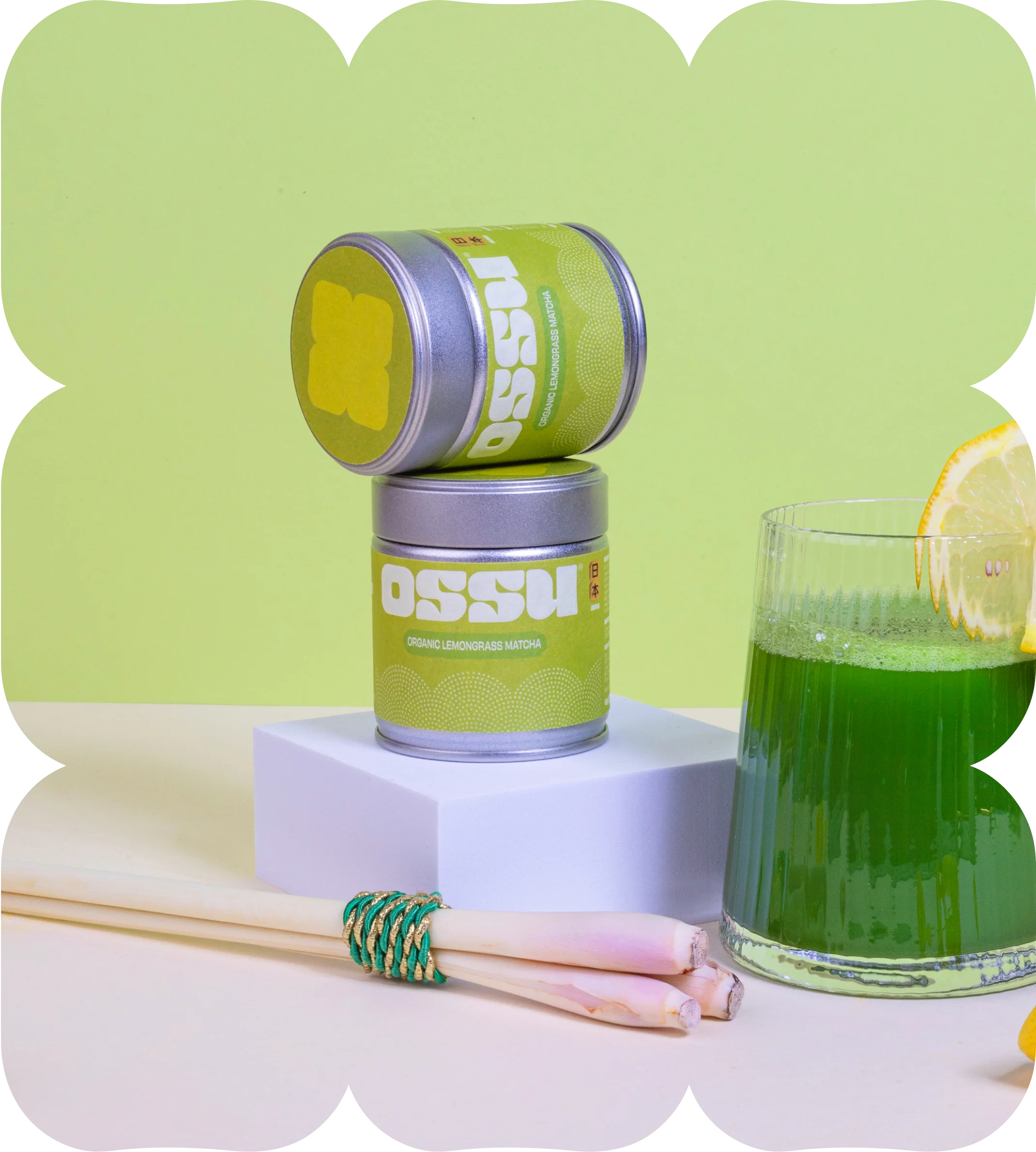 Two tins of OSSU organic lemongrass matcha stacked beside a glass of matcha drink with fresh lemongrass stalks