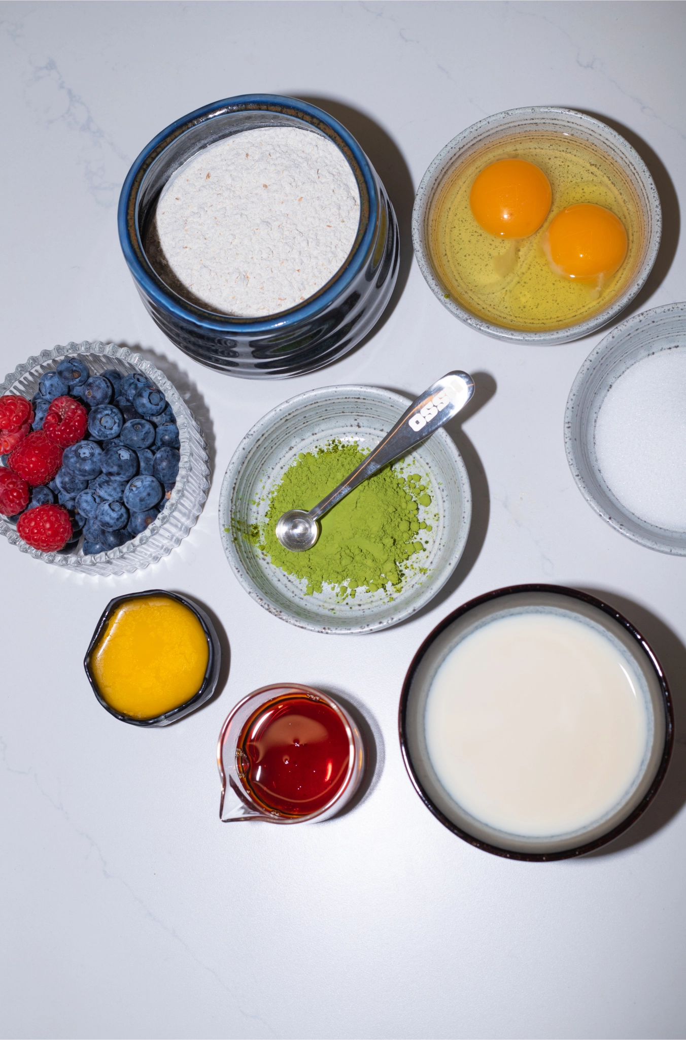 Baking ingredients arranged on a countertop including flour, eggs, berries, matcha powder, milk, honey, and sugar in small bowls
