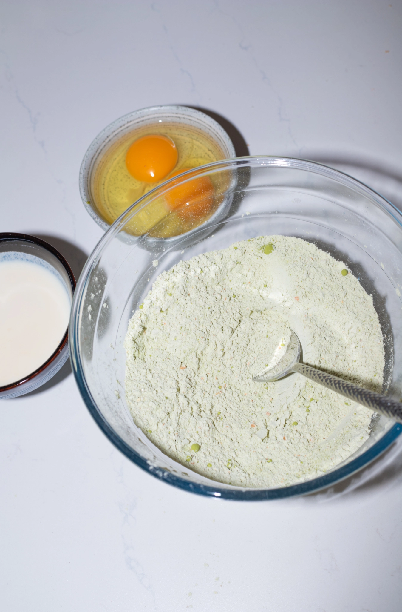 Flour mixture in a glass bowl with a spoon beside bowls of milk and cracked eggs on a countertop