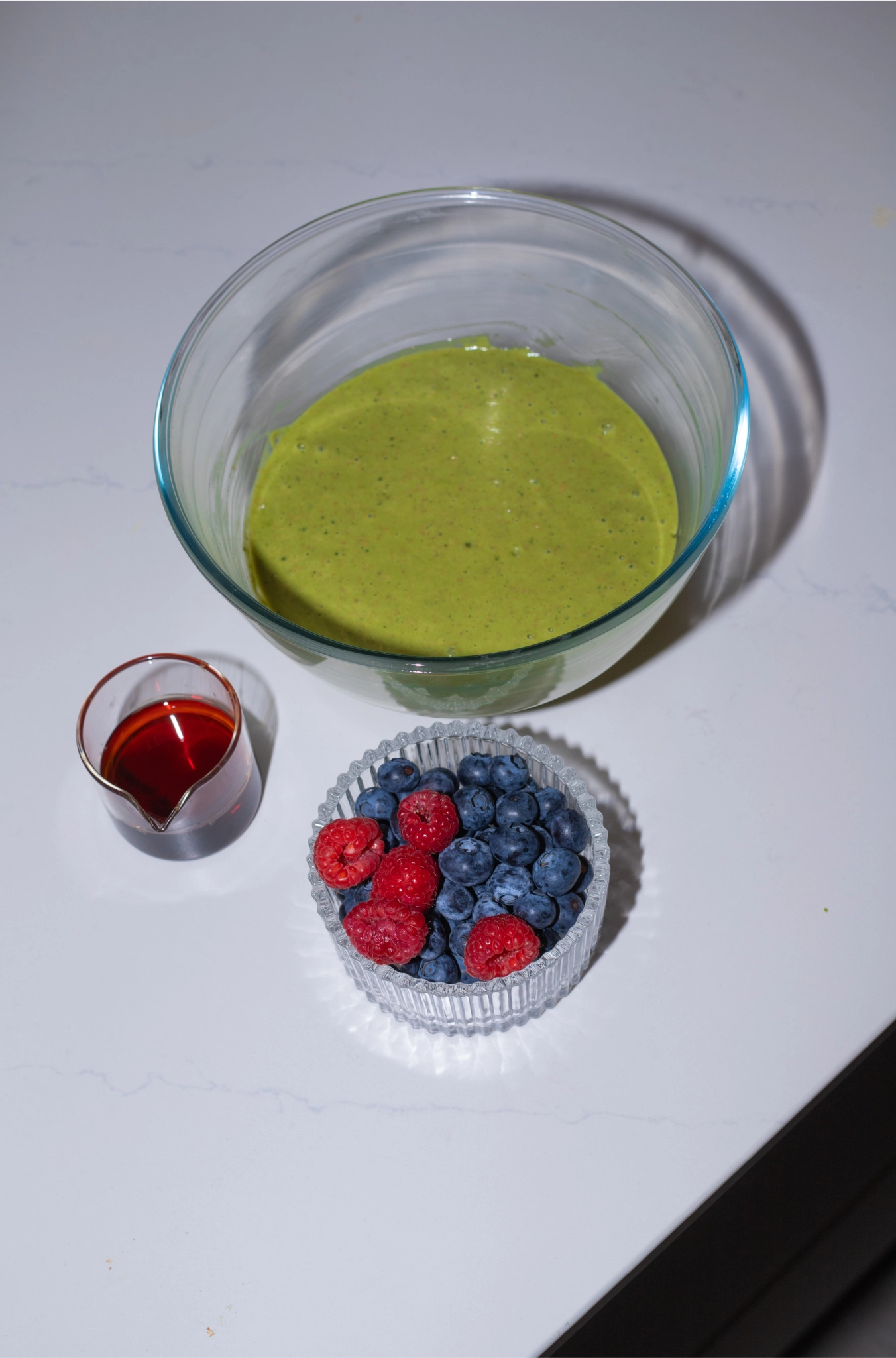 Bowl of green matcha batter with a small pitcher of syrup and a dish of fresh blueberries and raspberries on a countertop