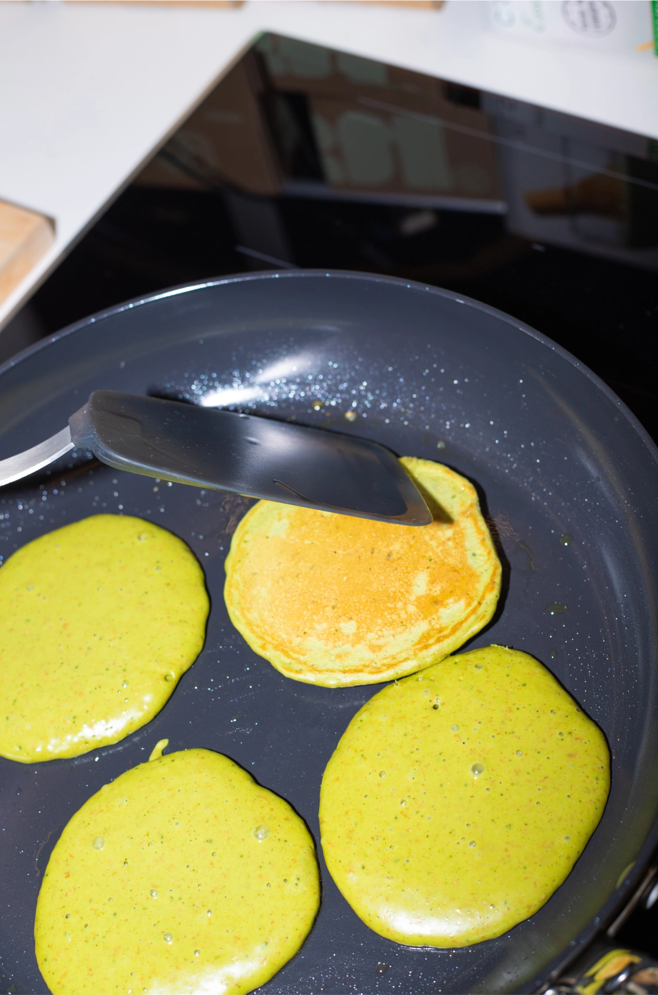 Matcha pancakes cooking in a frying pan, with a spatula flipping one golden pancake