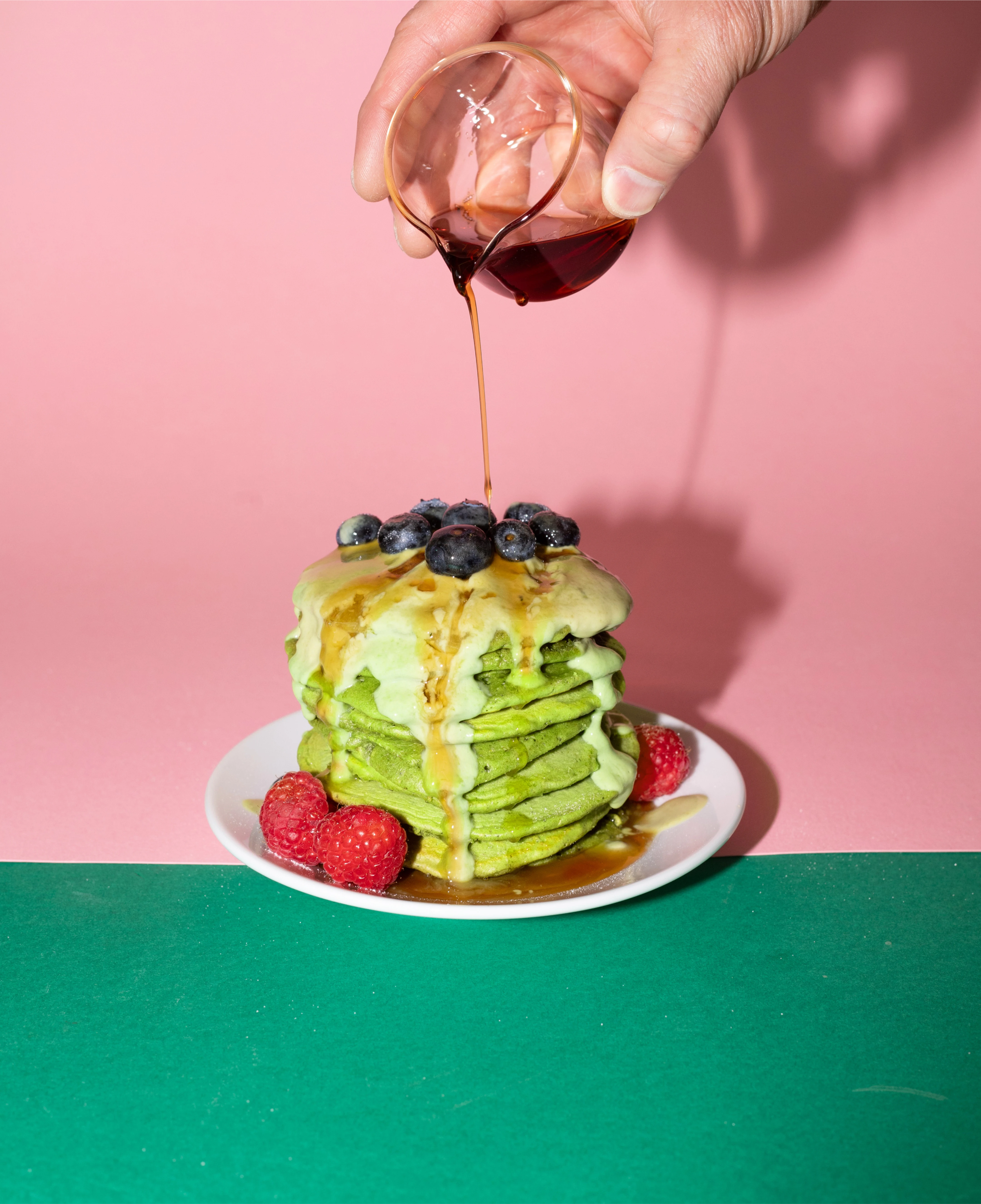Stack of green matcha pancakes topped with blueberries and raspberries as syrup is poured over