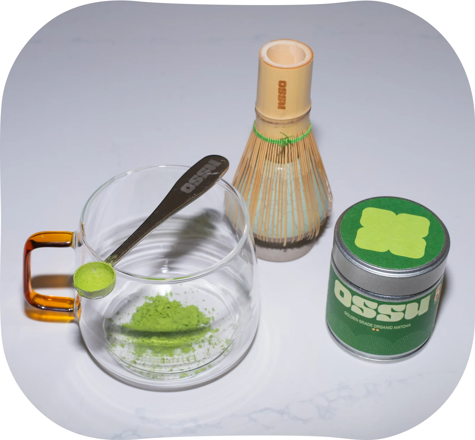 Matcha powder being added to a glass cup with a bamboo whisk and OSSU matcha tin beside it