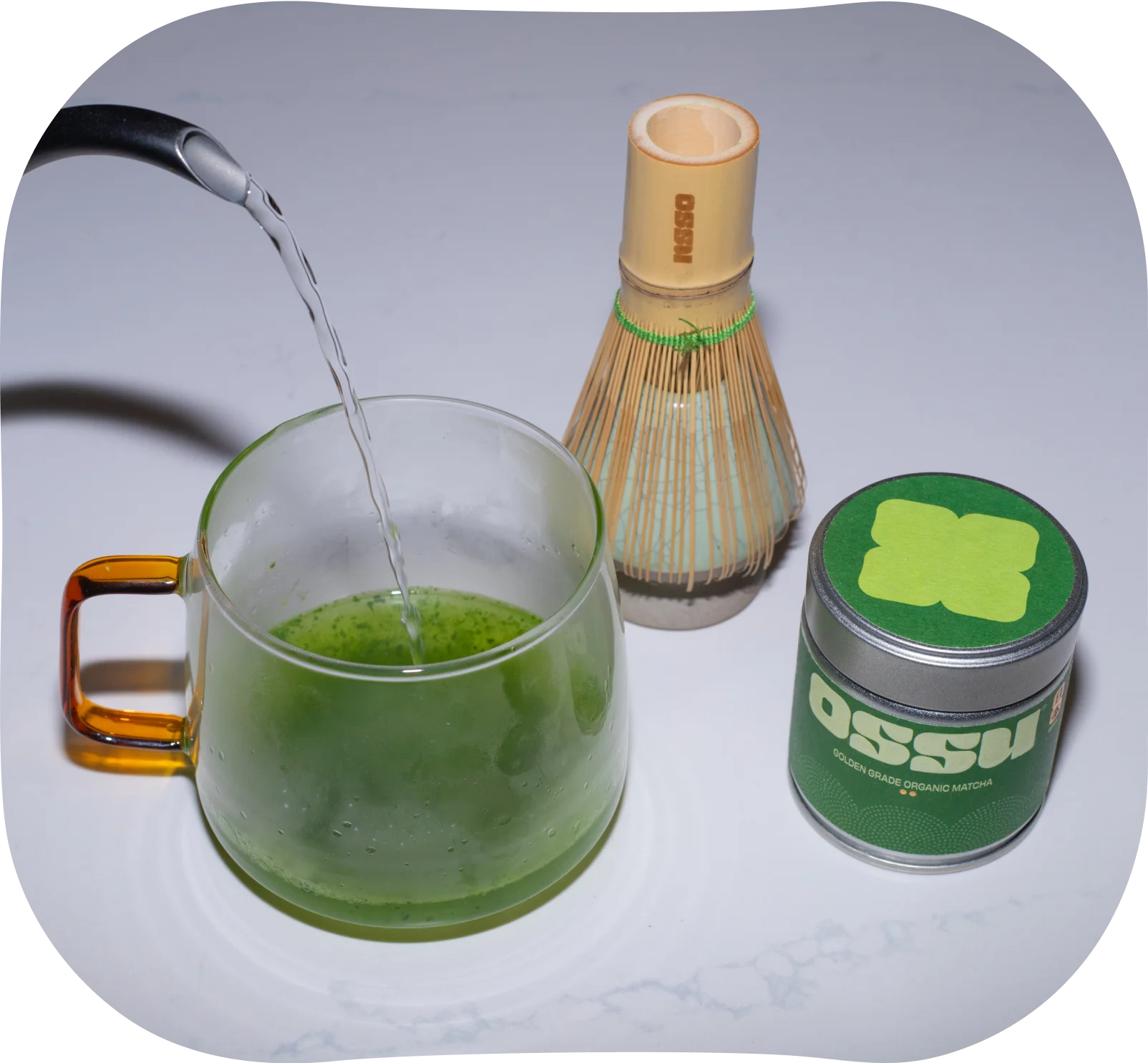 Hot water being poured onto matcha powder in a glass cup with bamboo whisk and OSSU matcha tin nearby