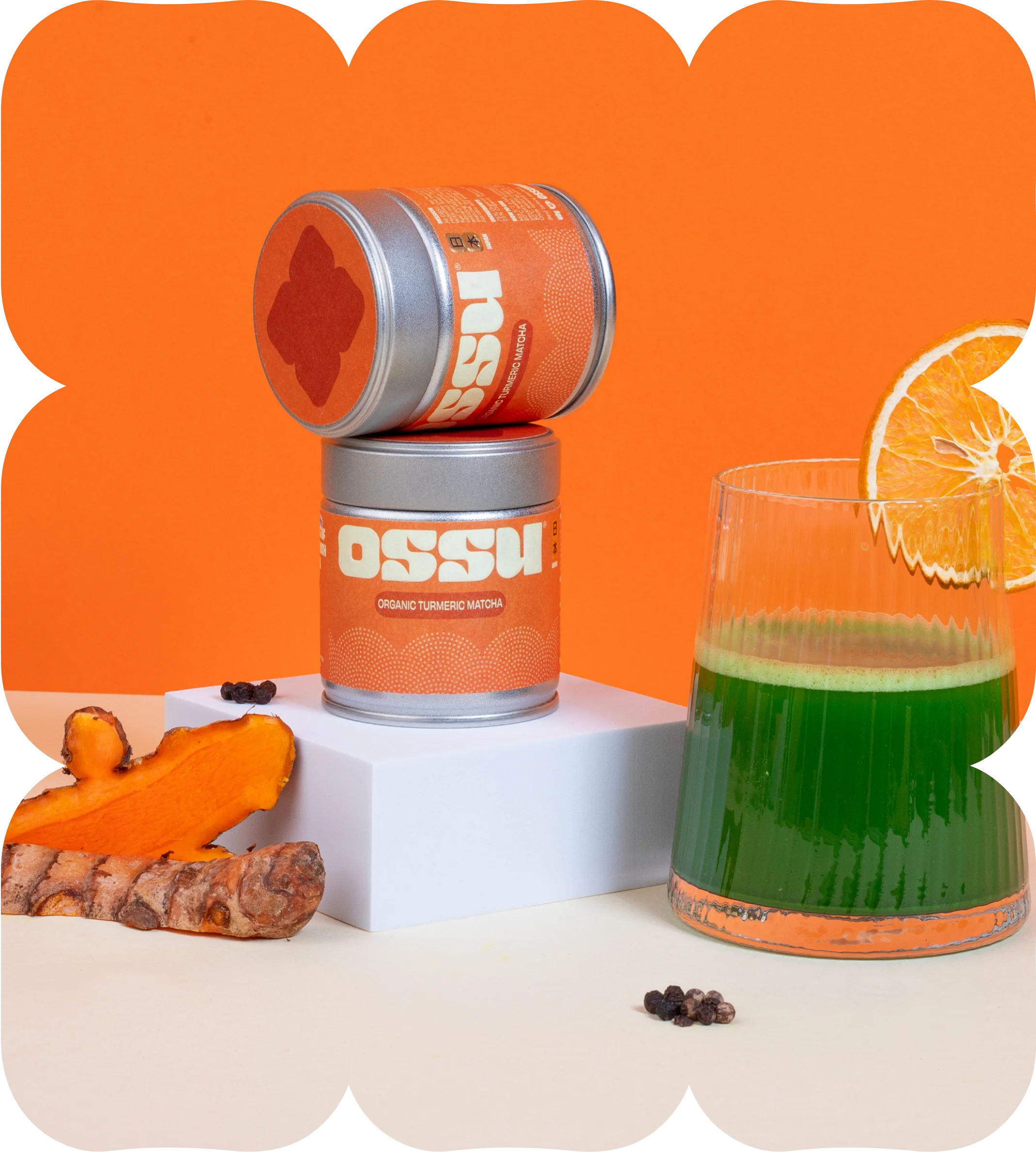 Two tins of OSSU organic turmeric matcha stacked beside fresh turmeric root and a glass of green matcha drink with an orange slice
