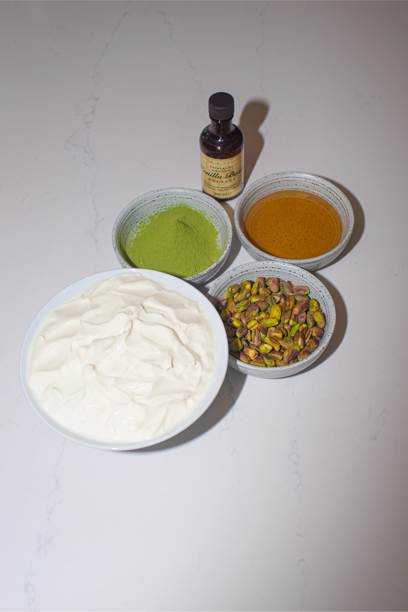 Bowls of yogurt, matcha powder, honey, and pistachios arranged on a countertop with a small bottle of vanilla extract
