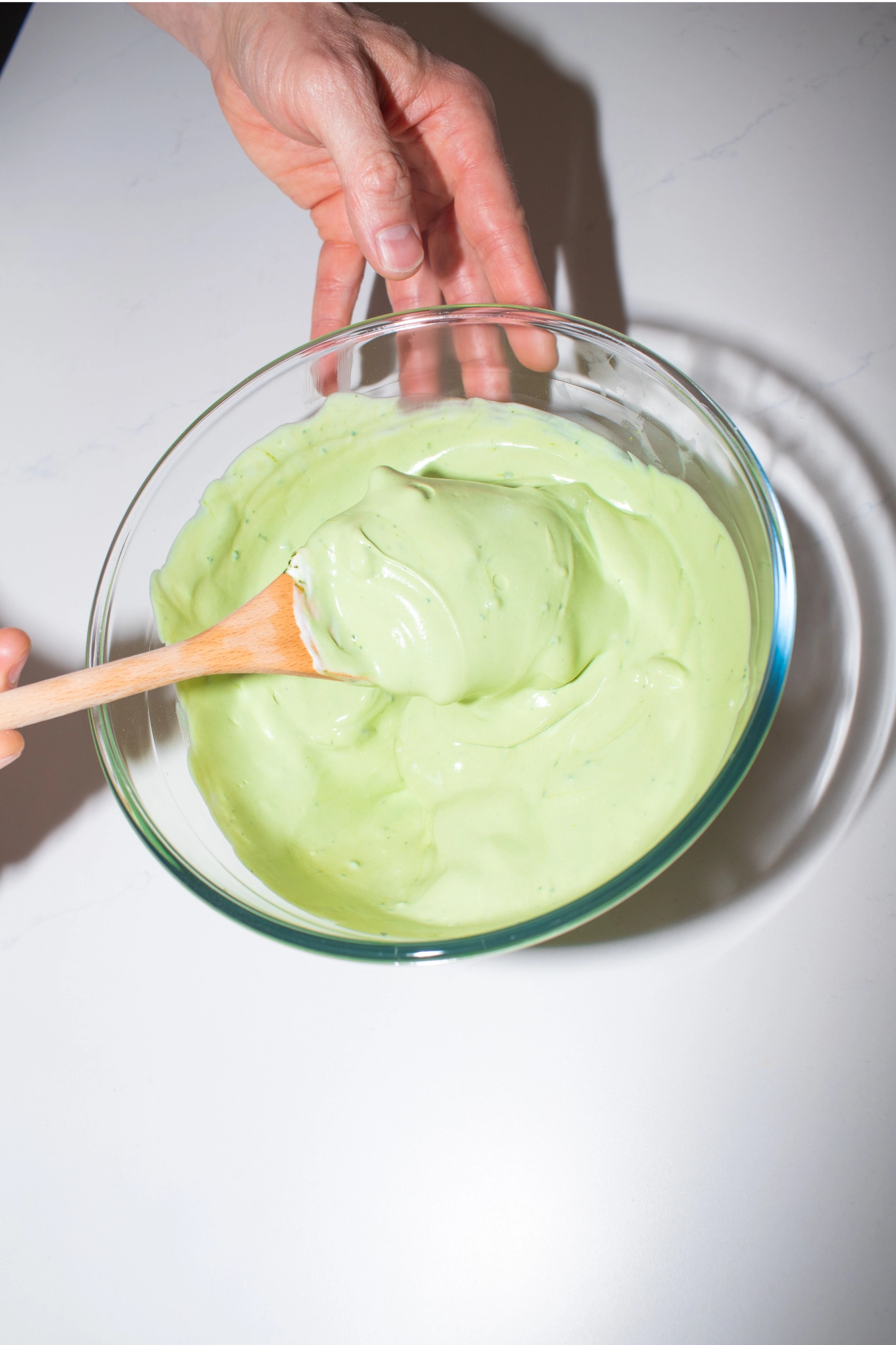 Hand stirring smooth pale green matcha yoghurt mix in a glass mixing bowl with a wooden spatula