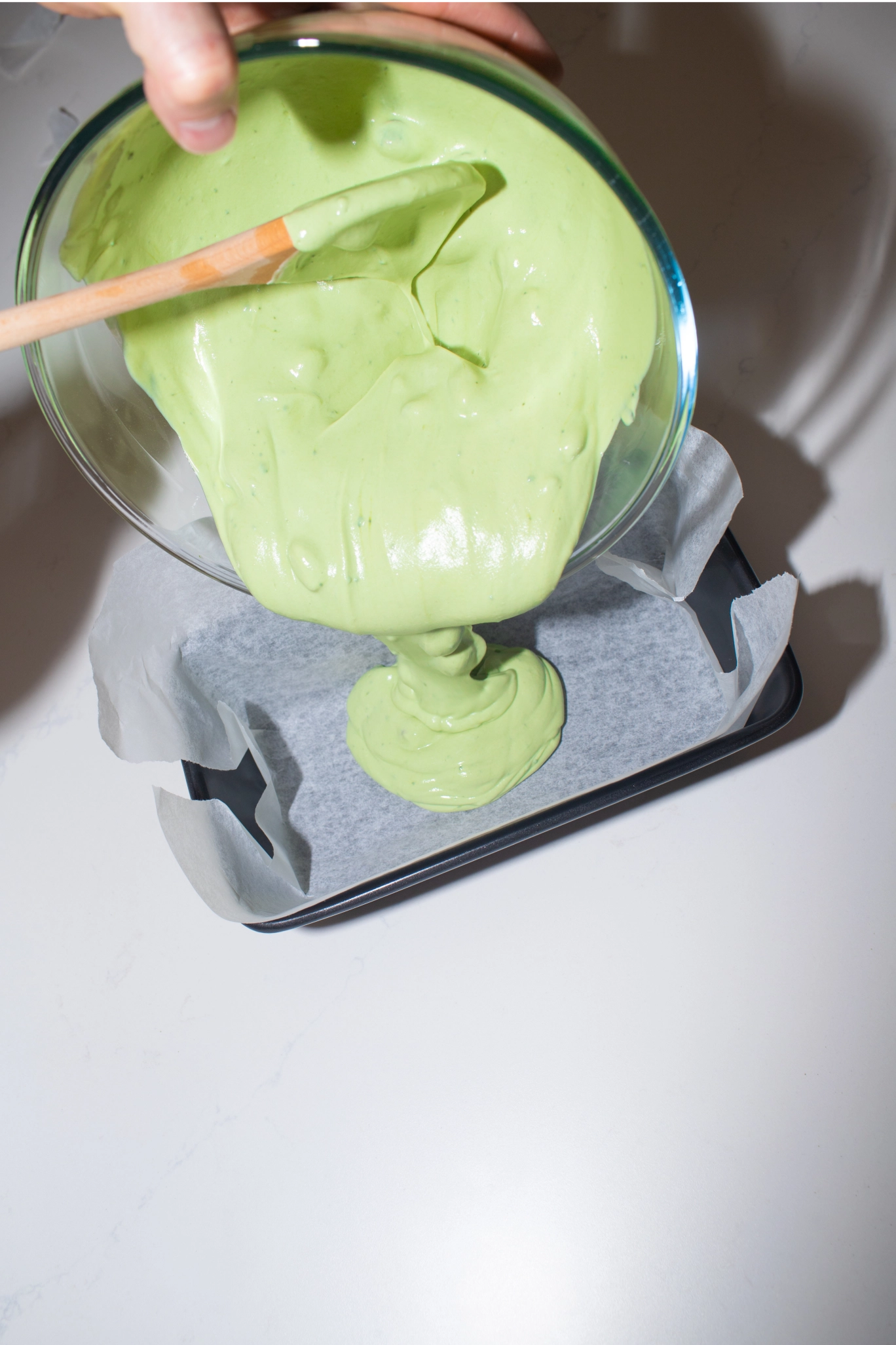 Pouring thick pale green matcha yoghurt mix from a glass bowl into a parchment-lined baking tray