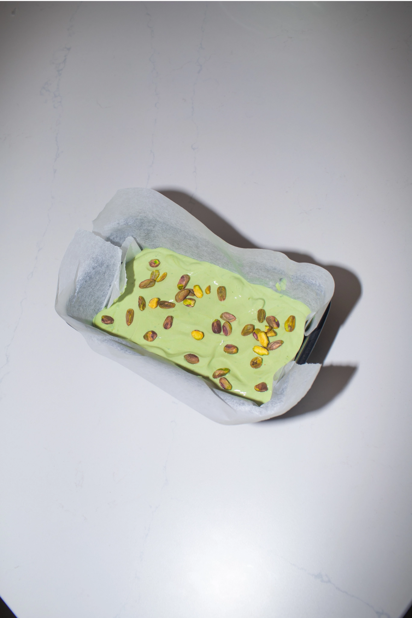 Matcha frozen yoghurt mixture in a parchment-lined tray topped with chopped pistachios