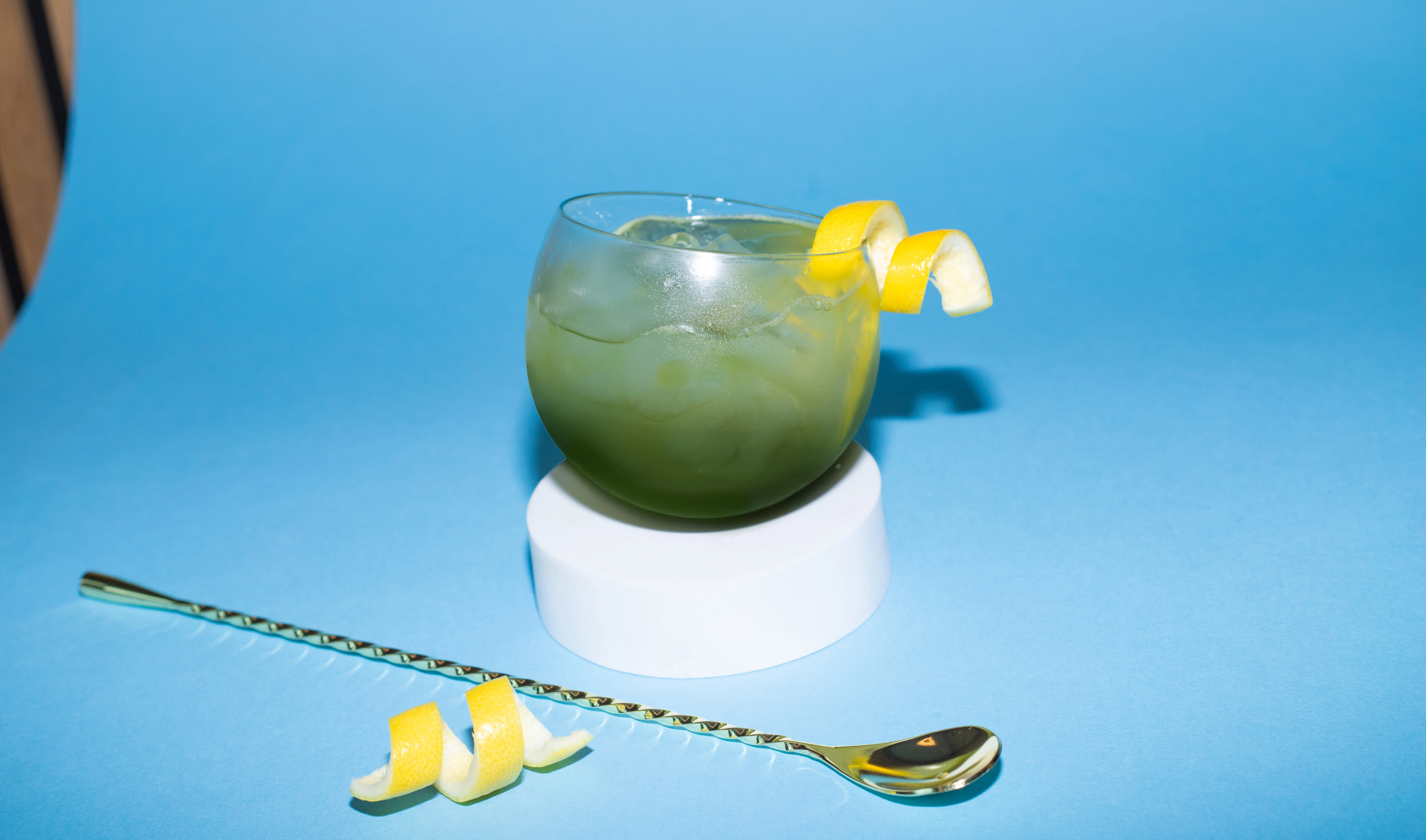 Glass of whiskey iced matcha with lemon garnish on a blue background, resting on a white stand with a spoon beside it