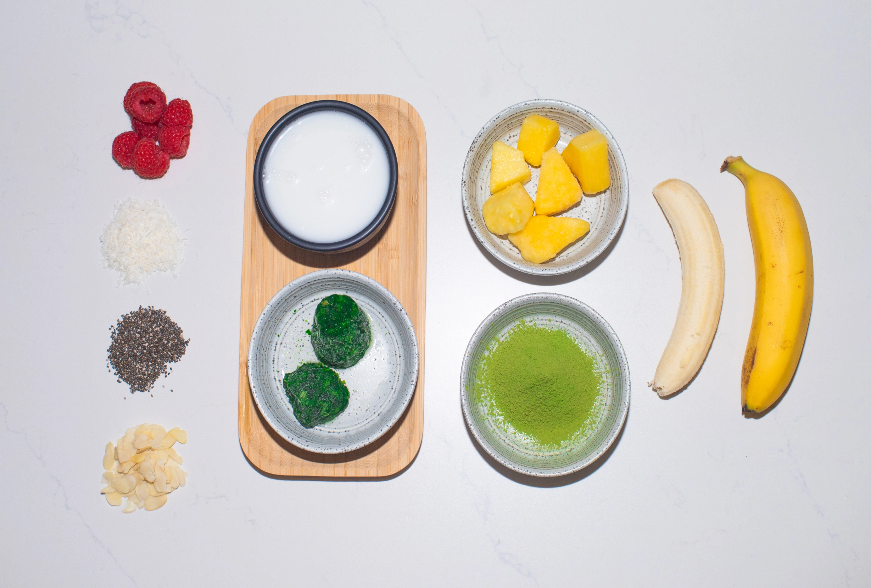 Flat lay of smoothie ingredients including matcha powder, frozen green cubes, coconut milk, pineapple chunks, banana, raspberries, chia seeds, coconut flakes, and sliced almonds arranged on a white surface