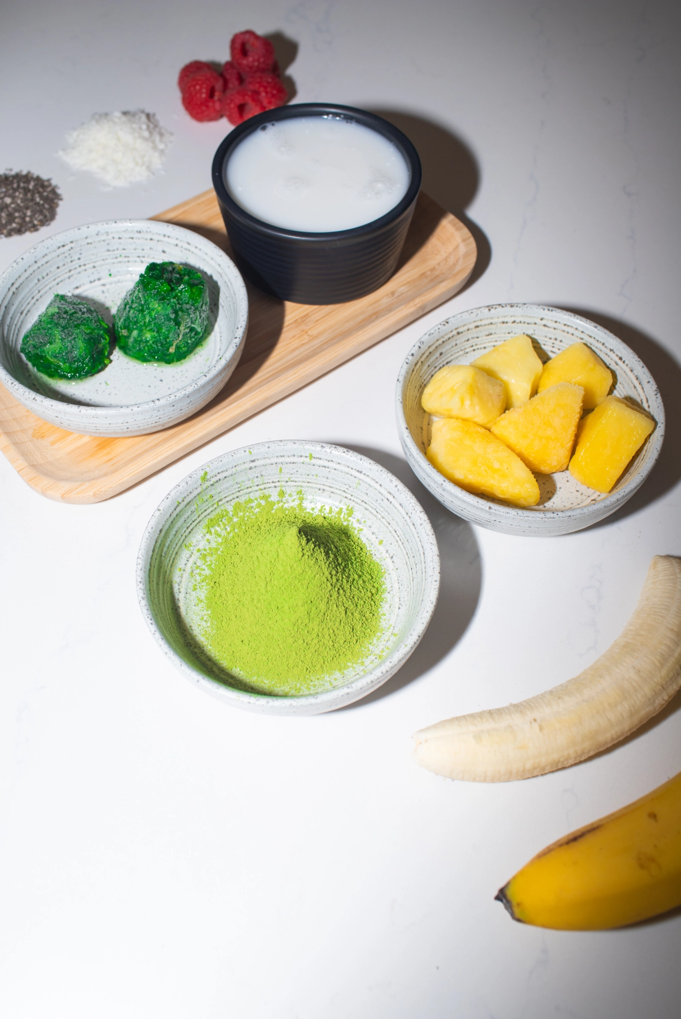 Top-down view of matcha smoothie ingredients including matcha powder, frozen greens, coconut milk, pineapple chunks, banana, raspberries, seeds, and coconut arranged on a countertop