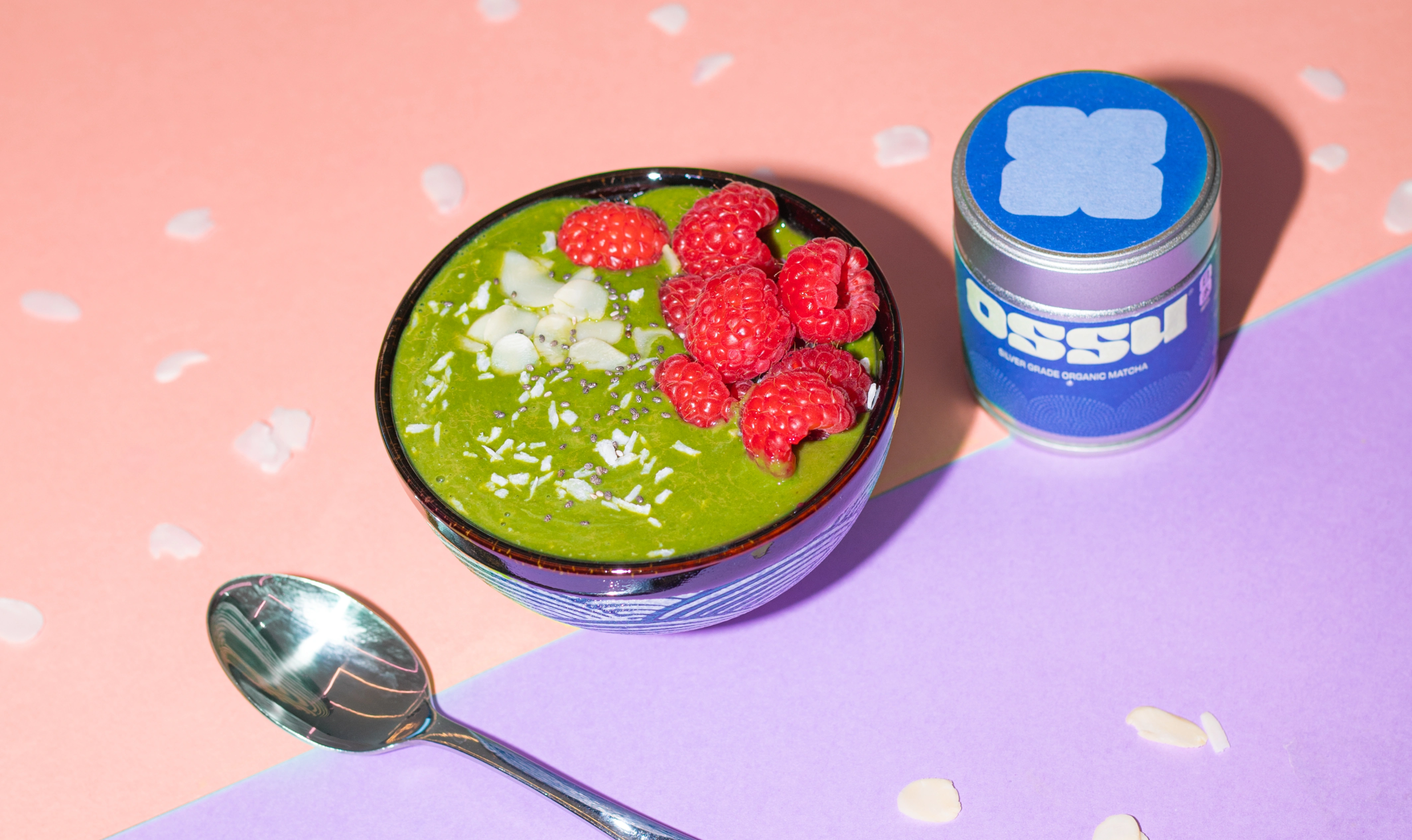 Bowl of green matcha smoothie topped with raspberries and coconut flakes beside a tin of OSSU matcha on a colourful surface